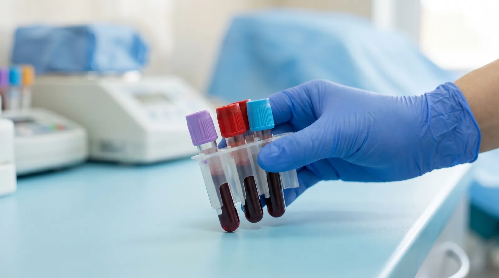 A gloved phlebotomist's hand holding a vacutainer needle holder with several color-capped collection tubes filling with dark red blood, on a clean clinical surface in soft blue light.